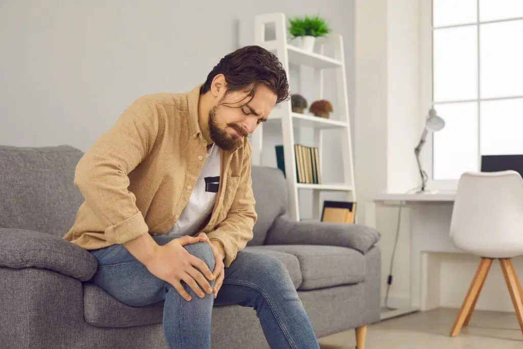 Man sitting on a couch because of knee pain bends over and holds his knee while making a face.