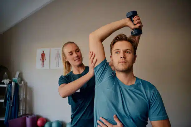 Young physiotherapist helping man to do a physical therapy exercise with dumbbells.