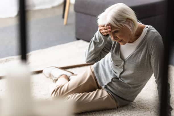 senior woman with vertigo sitting on carpet and touching forehead with hand feeling dizzy.